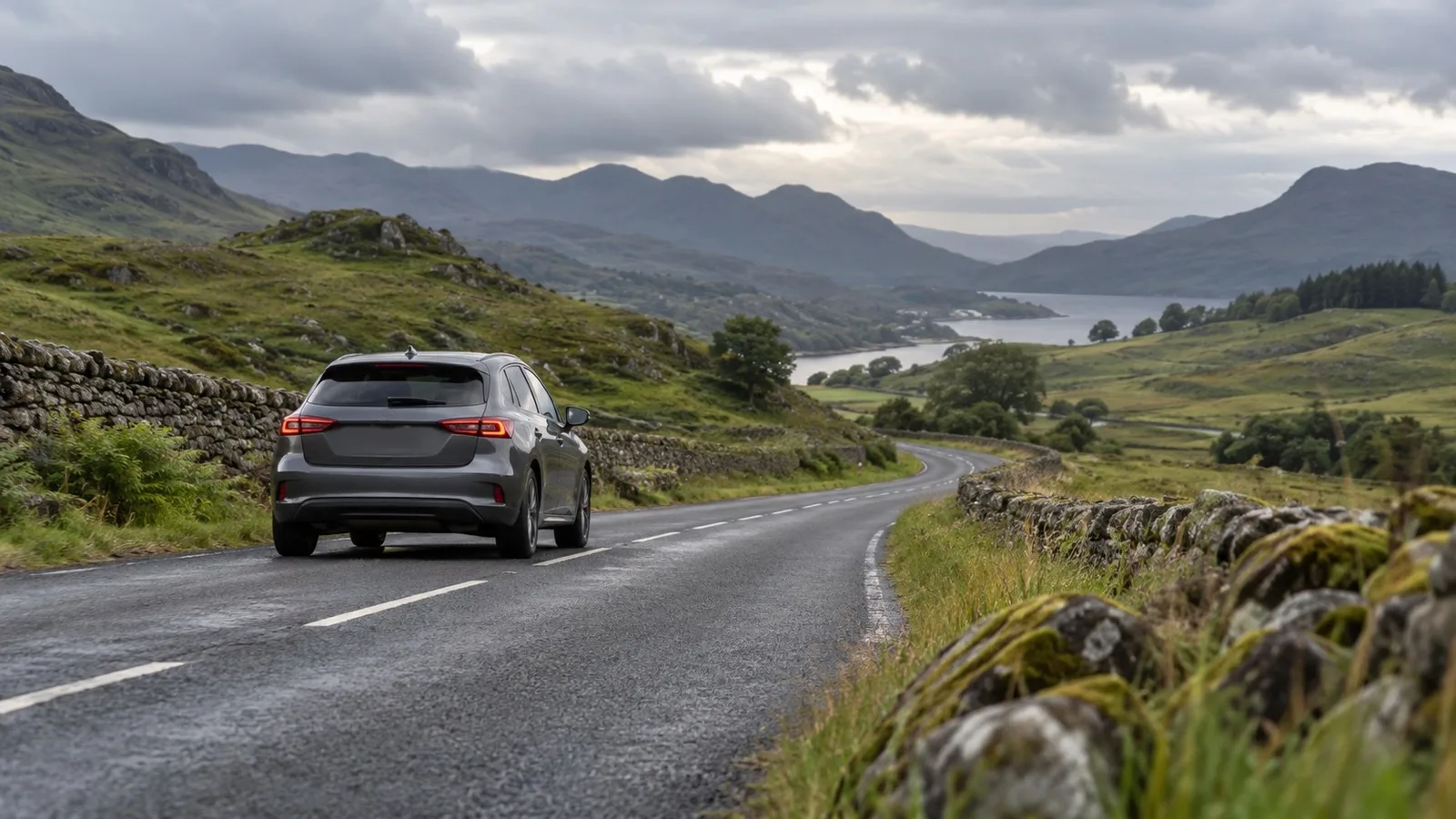 A car driving along a winding rural road through the Scottish Highlands, dry stone walls alongside a loch in the distance