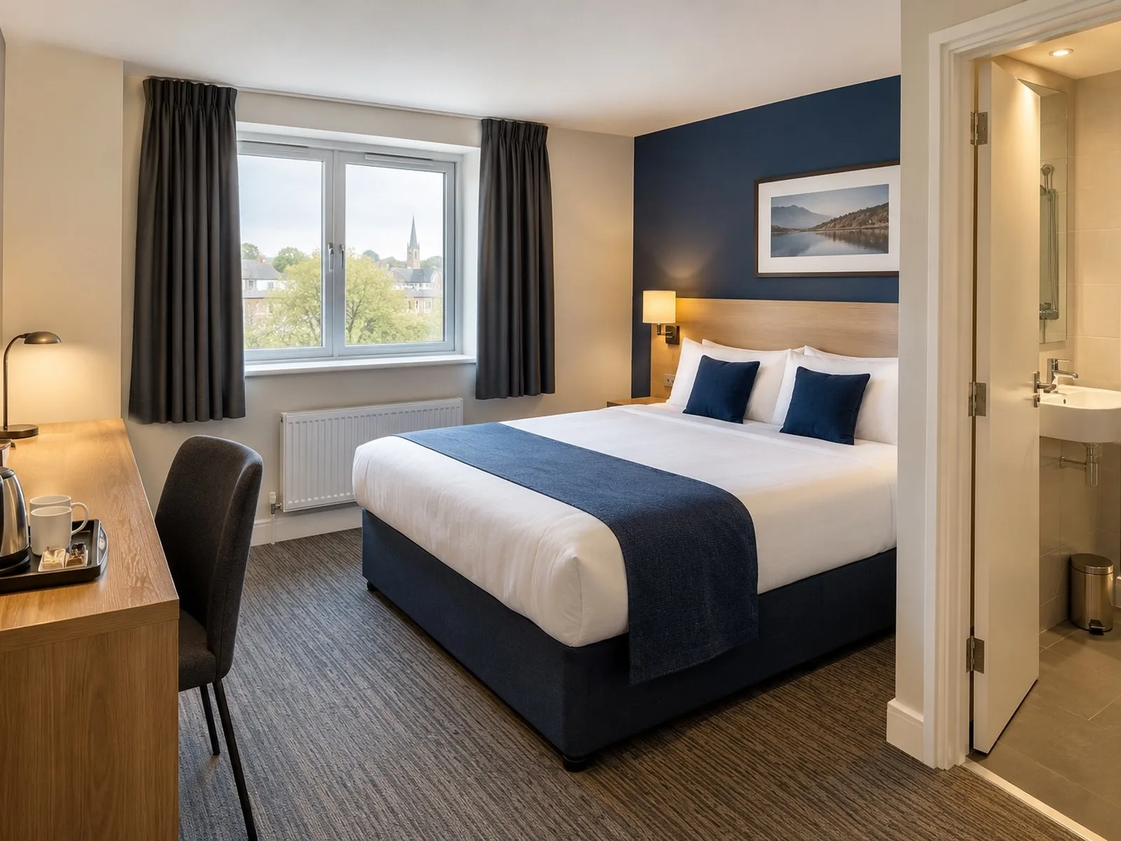 A clean modern budget hotel room with a navy-and-white bed, work desk and en-suite bathroom visible through an open door